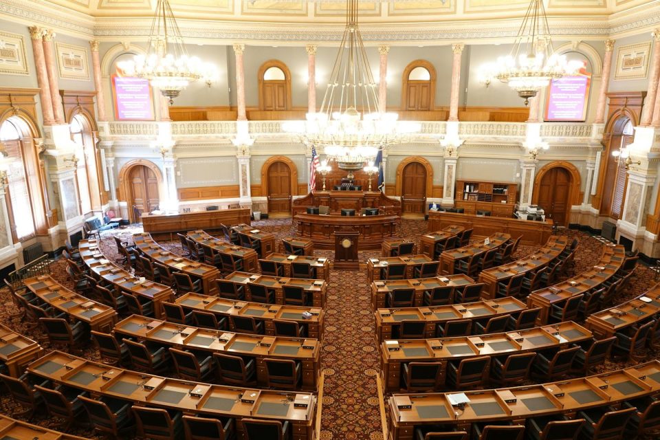 Wide view of an ornate legislative chamber with empty seats and chandeliers.