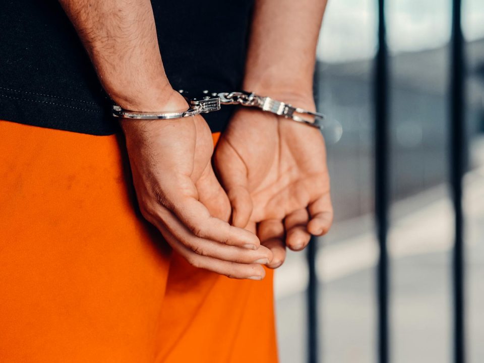 Close-up of person in handcuffs wearing orange attire behind prison bars, symbolizing arrest.