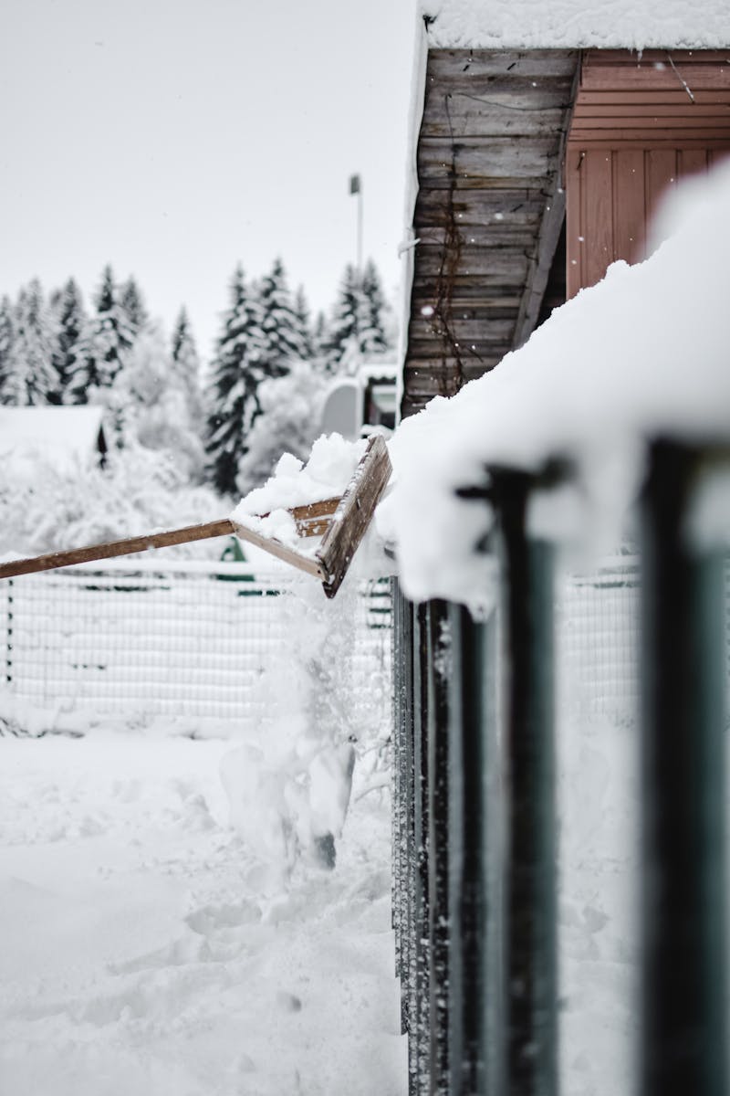 A winter scene showing snow being pushed off a fence with trees and buildings in the background.