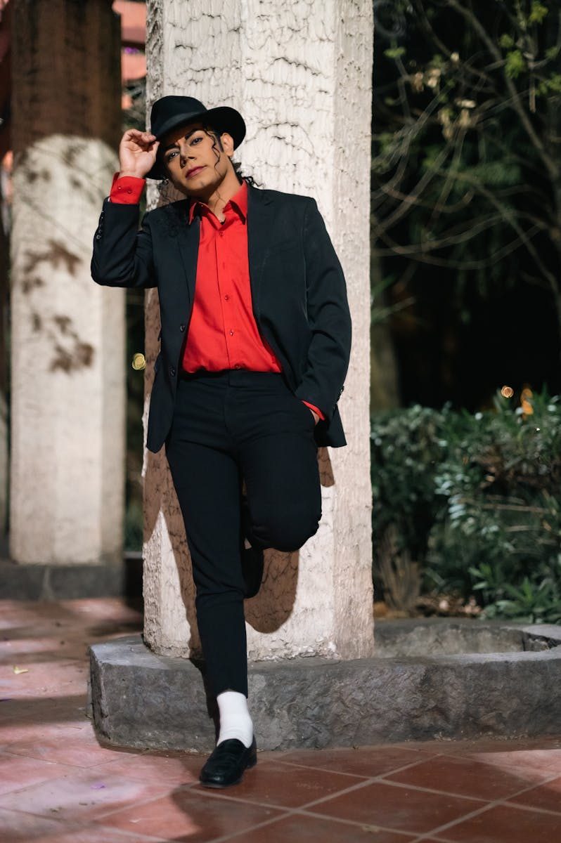 Fashionable man in a red shirt and black suit wearing a hat, leaning against a pillar outdoors.