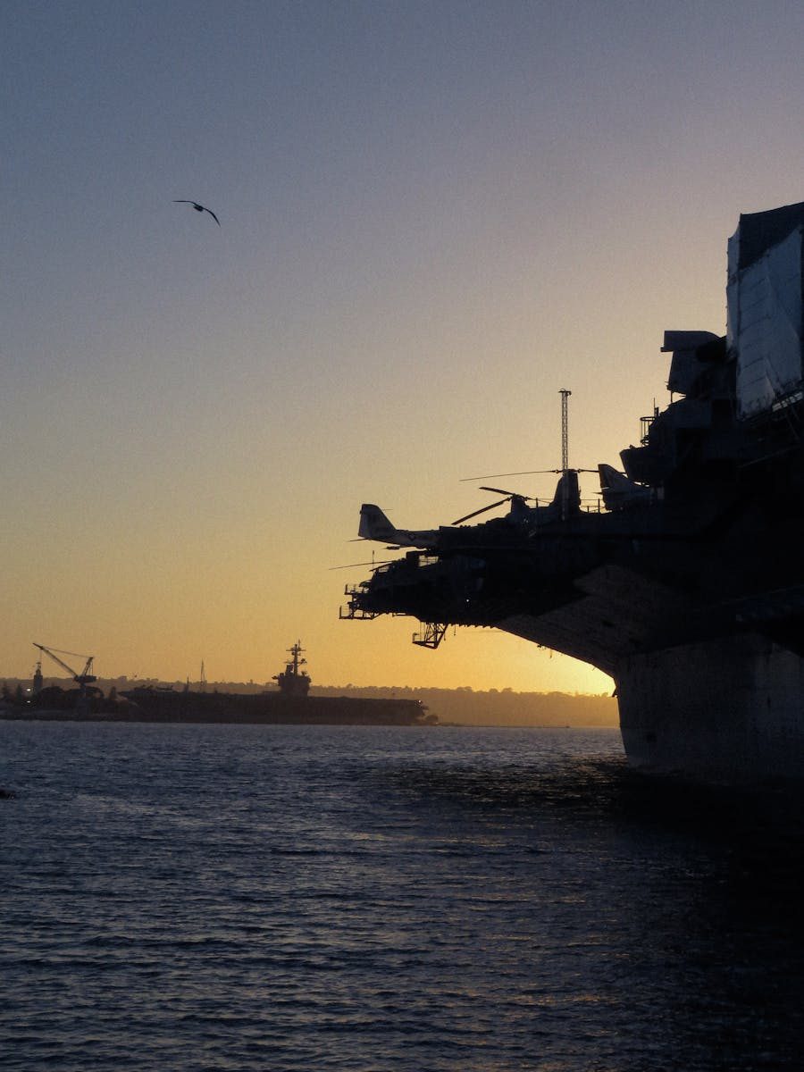 Silhouette of an aircraft carrier at sunset in a harbor, emphasizing military strength.