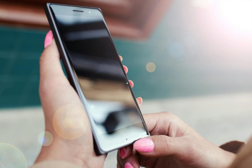 Close-up of hands holding a smartphone outdoors with a bright lens flare and reflections.
