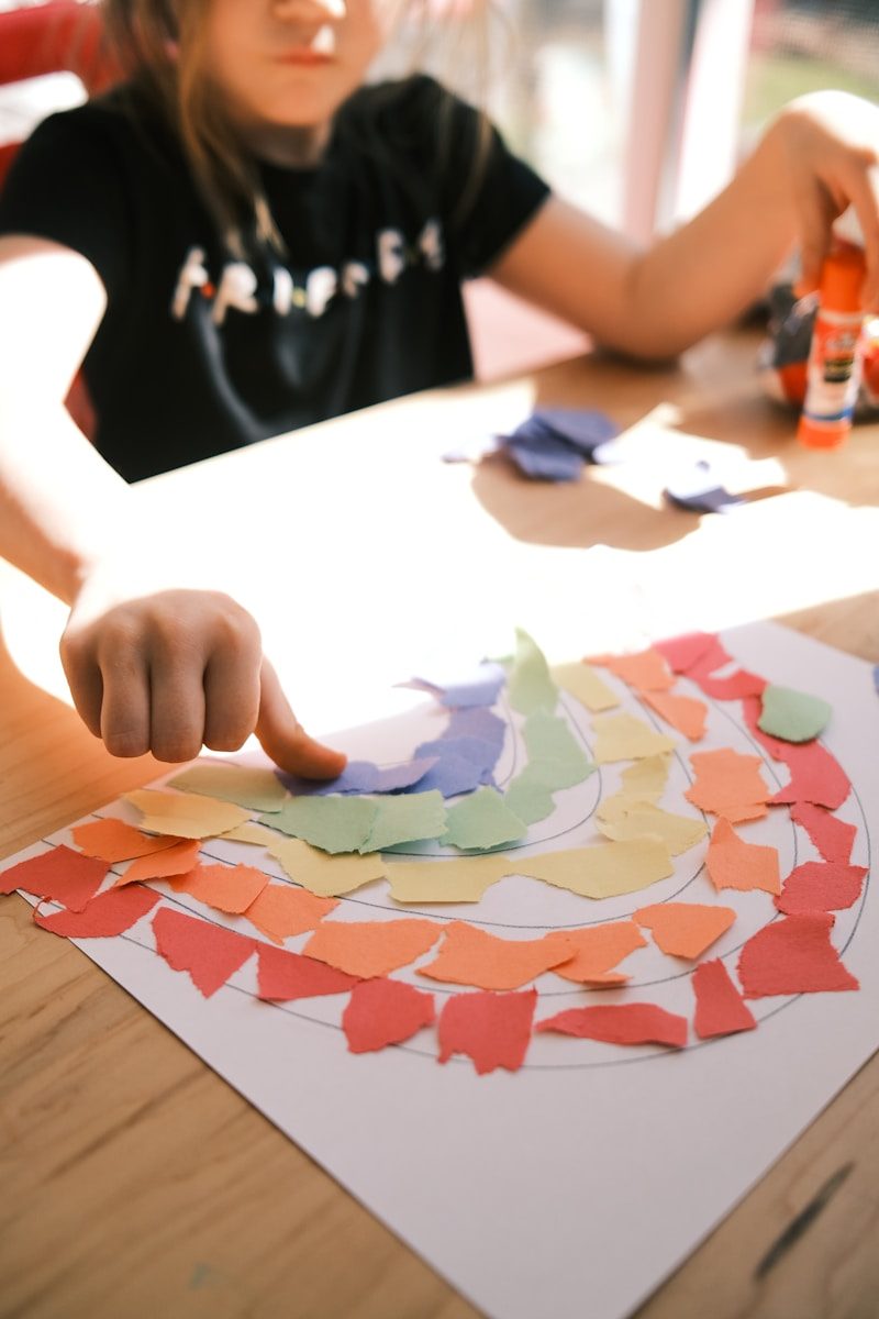 A child gluing colorful paper scraps onto a drawing of a rainbow