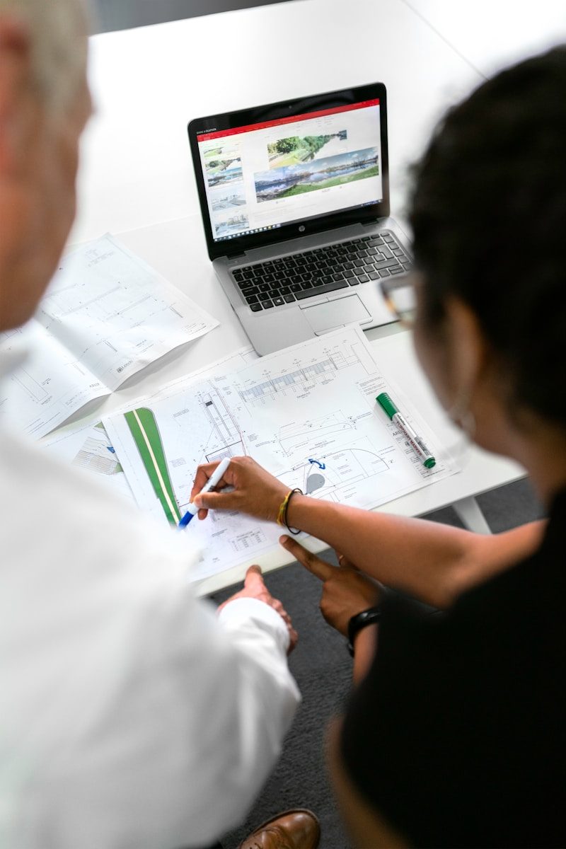 Two people reviewing architectural blueprints on a desk with a laptop in a workspace